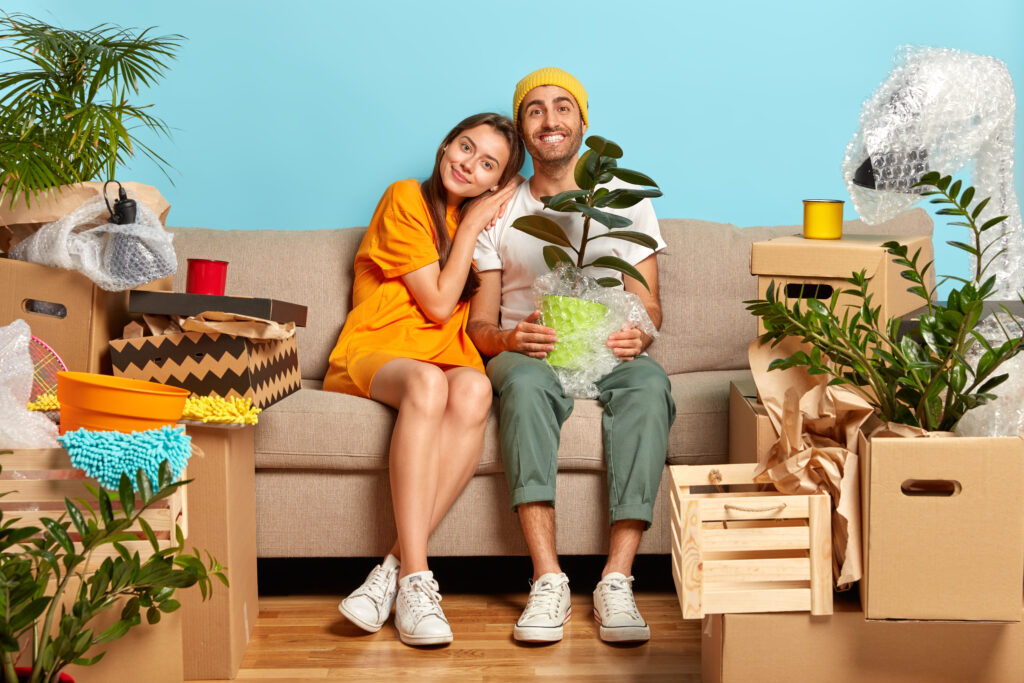 smiling couple sitting on a couch surrounded by moving boxes and houseplants